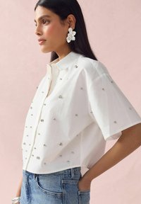 Chemise blanche à boutons ornée de broderies en strass éparpillées, à manches courtes et à coupe relaxée, associée à un jean bleu clair.