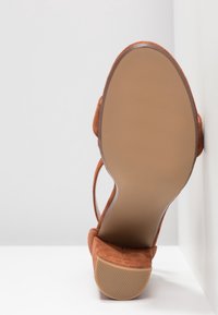Sandale en daim marron avec un talon carré vue du dessous, positionnée contre un fond blanc en coin.