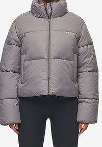 Grå puffjakke med høj krave, frontlynlås og logo på brystet, båret over sorte tætsiddende leggings.
