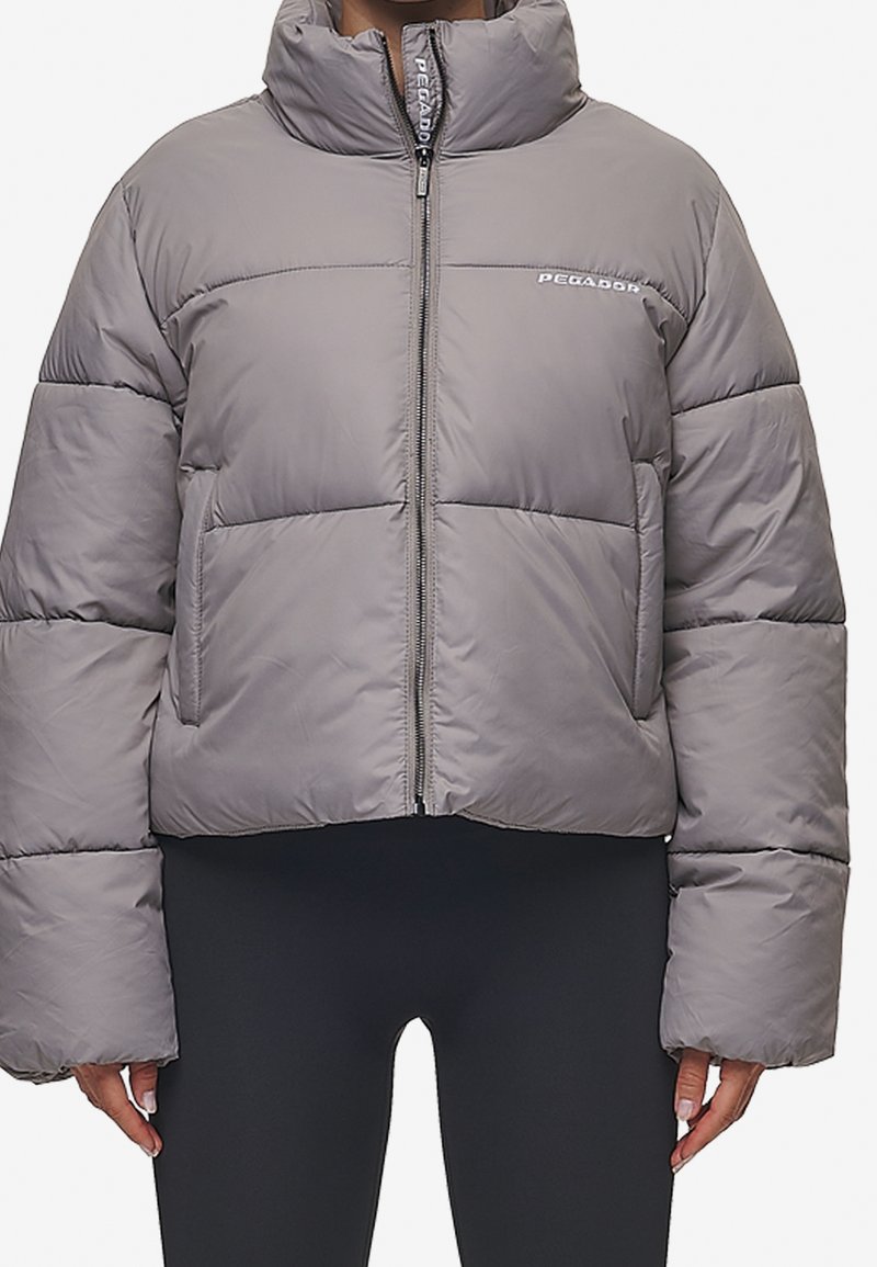Grå puffjakke med høj krave, frontlynlås og logo på brystet, båret over sorte tætsiddende leggings.