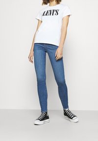 Camiseta blanca con el texto "LEVI'S", jeans ajustados azules y zapatillas negras de caña alta. Diseño simple, corte casual y texturas de color sólido.