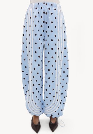 Pantalon large bleu clair à pois noirs, bandes blanches incurvées sur les côtés, ceinture élastique, porté avec des chaussures noires pointues.