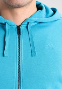 Felpa con zip blu realizzata in morbido tessuto, dotata di tasca a marsupio, con corde regolabili e logo Adidas in rilievo.