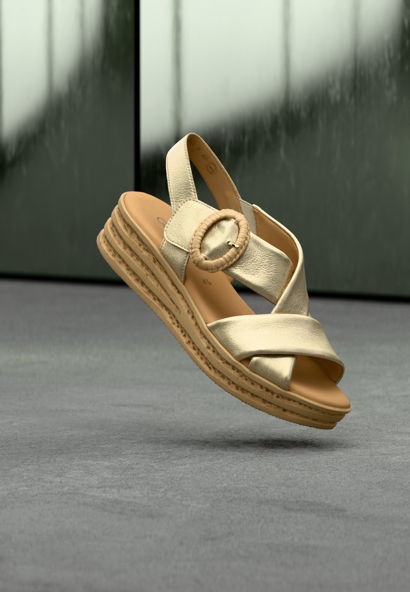 Sandale à plateforme en cuir beige avec des brides croisées, une boucle ronde et une semelle texturée flottant au-dessus d'une surface grise.