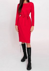 Femme portant une robe rouge mi-longue à manches longues avec une ceinture à la taille, associée à des bottes noires lacées montantes.