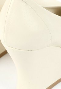 Gros plan sur la zone du talon d'une chaussure à talons hauts en cuir crème avec des détails de couture visibles.