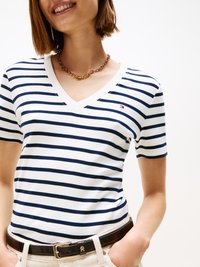 Camiseta branca de gola em V com riscas horizontais em azul marinho, apresentando um pequeno logótipo no peito. Combinada com um cinto escuro e um colar de corrente.