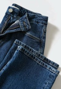 Vaqueros de mezclilla azul con cremallera, diseño de cinco bolsillos, costuras visibles en un dorado contrastante y una textura ligeramente descolorida en la tela.