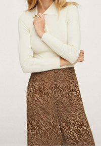 Top blanco de manga larga con cuello, combinado con pantalones marrones con un patrón de lunares. El conjunto presenta materiales ajustados y texturizados.