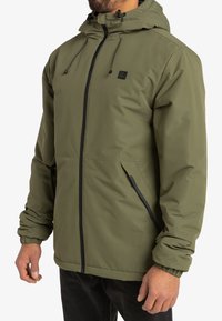 Uomo che indossa una giacca a cappuccio verde oliva con zip frontale, polsini elasticizzati e tasche laterali con zip angolata, in piedi contro uno sfondo bianco.