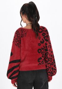 Maglione rosso, peloso, con stampa leopardata nera e motivi astratti. Design con spalle scese e tessuto texturizzato, abbinato a pantaloni scuri.