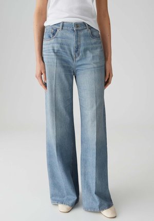 Person trägt hellblaue Jeans mit weitem Bein und Frontfalten, weißes Oberteil und weiße Schuhe, steht vor einem schlichten hellen Hintergrund.