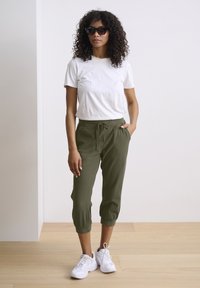 Vit bomullst-shirt, lösa olivgröna korta byxor med dragsko och vita sportskor. Modellen bär mörka solglasögon. Mjuk textur.