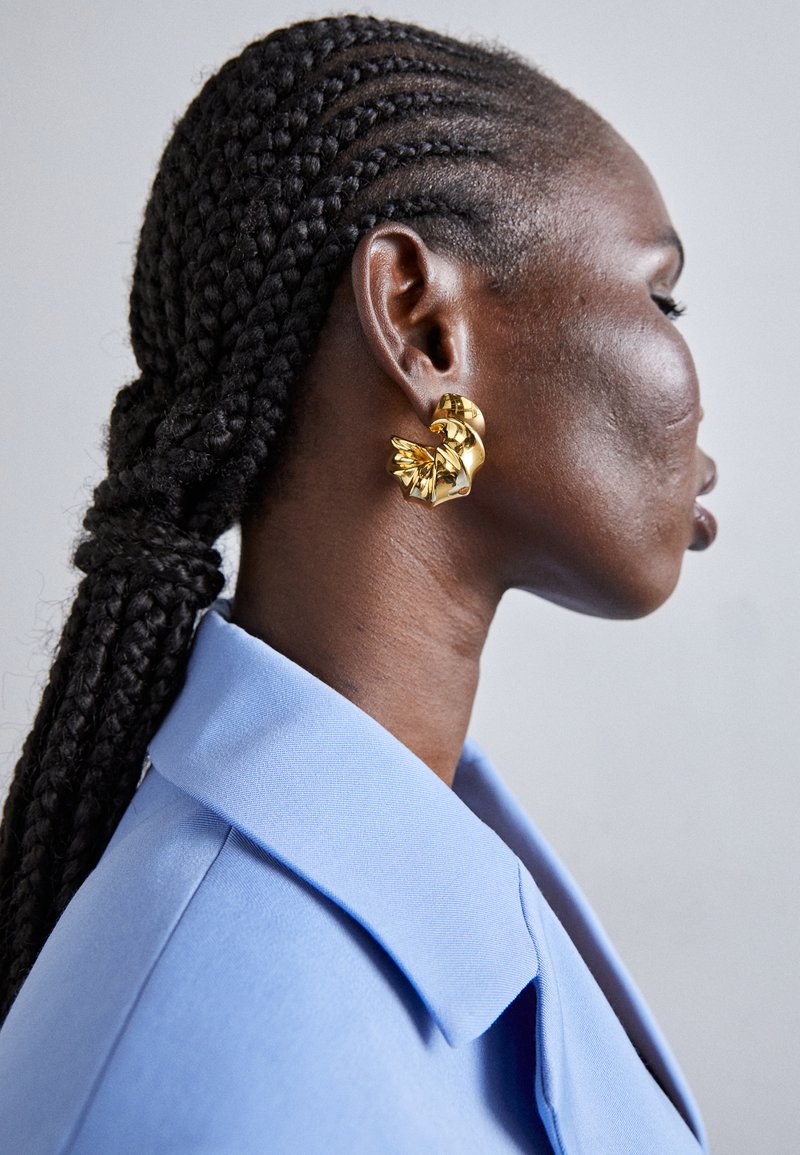 Pendientes de declaración en oro con un diseño escultórico, que presentan formas en espiral y superficies lisas, llevados por una persona con cabello trenzado.