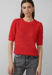 Rode, gestructureerde gebreide trui met korte mouwen en een ronde hals, gecombineerd met grijze denim jeans. De stof lijkt zacht en een beetje pluizig.