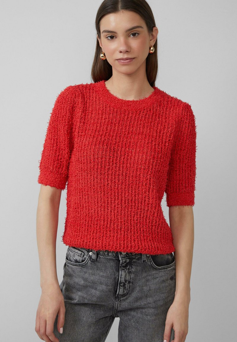 Rode, gestructureerde gebreide trui met korte mouwen en een ronde hals, gecombineerd met grijze denim jeans. De stof lijkt zacht en een beetje pluizig.