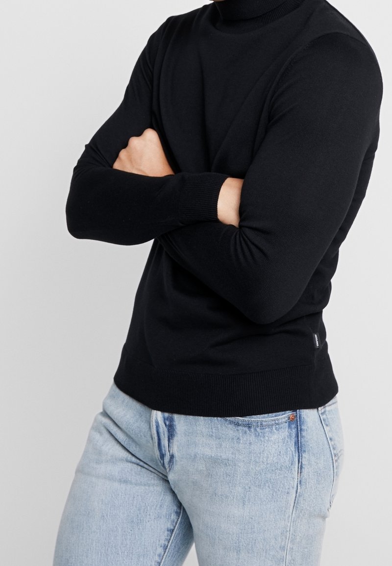 Schwarzer Strickpullover mit hohem Kragen, eng anliegenden Ärmeln und geripptem Saum, kombiniert mit hellblauen Jeans. Keine sichtbaren Muster oder Beschläge.