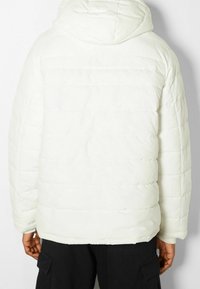 Vit vadderad jacka med huva, med ett quiltat mönster och elastiska ärmslut. Slät struktur, lätt design och något oversized passform.