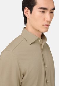 Camisa de botón color tostado con cuello clásico, textura suave y un diseño ajustado. Presenta un dobladillo redondeado y detalles de costura sutiles.