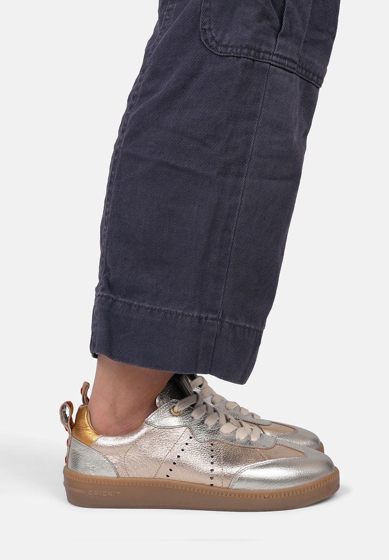 Metallic silberne Sneakers mit braunen Gummisohlen, runden Schnürsenkeln, perforiertem Design und einer goldenen Akzentuierung an der Ferse.