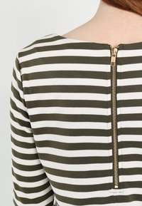 Haut à rayures vert olive et blanc avec un col rond et une fermeture éclair dorée à l'arrière. Tissu lisse, coupe ajustée et coutures nettes.