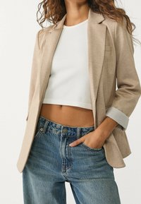 Blazer beige su misura con maniche leggermente svasate, indossato sopra un crop top bianco a costine, abbinato a jeans in denim a vita alta.