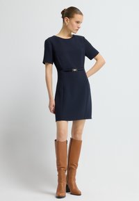Marineblauwe jurk met korte mouwen en een aansluitende silhouet, voorzien van een gouden accentriem. Bruine lederen laarzen tot de knie met een stevige hak.