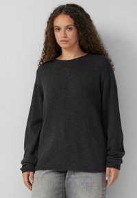 Vrouw met krullend haar, gekleed in een donkergrijze gehaakte trui en lichtgrijze jeans, staande tegen een effen lichtgrijze achtergrond.