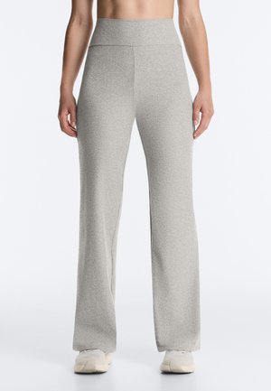 Femme portant un pantalon large taille haute gris clair avec des baskets blanches, debout devant un fond clair uni.