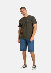 Camiseta de algodón verde oscuro de manga corta con un pequeño logo, combinada con pantalones cortos de mezclilla azul y zapatillas marrones con suelas blancas.