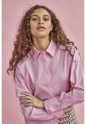 Vaaleanpunainen nappipaitapusero, jossa on kaulus, pitkät hihat ja sileä pinta, yhdistettynä kuvioituun hameeseen.