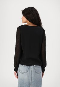Svart topp med gjennomsiktige, teksturerte ermer og rund hals, paret med en lett denimskjørt. Glatt stoff med subtile detaljer.