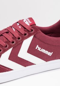 Bordowe sneakersy z białymi paskami w kształcie chevronu, białą podeszwą, bordowymi sznurówkami oraz logo "hummel" po bokach i na języku.
