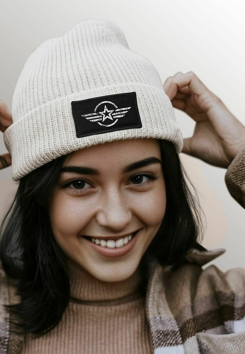 White ribbed beanie with a black rectangular patch featuring a star and wing design. The material appears soft and textured.