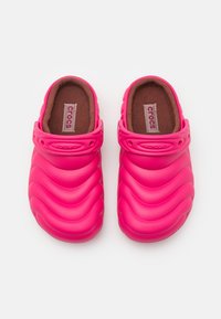 Crocs di un vivace colore rosa con un design ondulato e testurizzato, dotati di una soletta marrone imbottita e di una tracolla regolabile. Materiale leggero e flessibile.