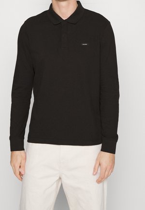 Homme portant un polo à manches longues noir avec un petit logo Calvin Klein sur la poitrine et un pantalon écru.