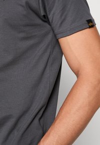 Šedé tričko s krátkým rukávem vyrobené z měkkého materiálu, které má jednoduchý design a štítek na rukávu pro branding.