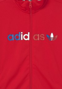 Červená zipová bunda s logem "adidas" v modré a bílé barvě, s ikonickými třemi pruhy v modré, bílé a červené barvě pod logem.