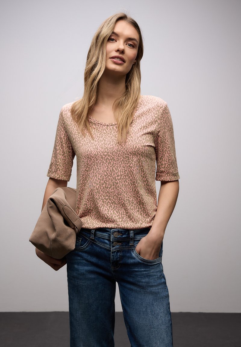 Pitkähiuksinen vaaleahiuksinen nainen, jolla on vaaleanpunainen leopardi-kuvioitu paita ja siniset farkut, pitäen toisessa kädessään taiteltua beigeä takkia.