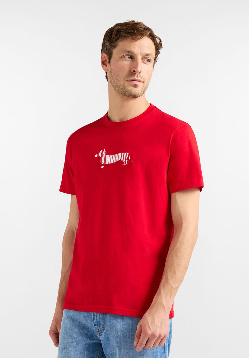 Camiseta de algodón roja con un gráfico de un dachshund a rayas blancas en el frente, cuello redondo y mangas cortas, combinada con jeans azules.