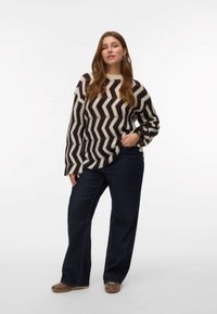 Pulover iz pletene tkanine s chevron vzorom v bežni in temno rjavi barvi, v kombinaciji s temnimi jeans hlačami s širokimi nogavicami. Pulover ima široke rokave in rebrast ovratnik.
