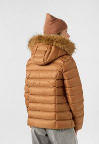 Bruine pufferjas met een capuchon gevoerd met bont, met een doorgestikt patroon en een getailleerde pasvorm. Gecombineerd met een zachte, tankleurige beanie en grijze jeans.