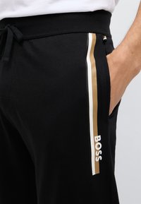 Czarne spodnie dresowe z regulowanym paskiem w talii na sznurek, bocznymi kieszeniami oraz kontrastowymi paskami w kolorze białym i złotym. Widoczne jest białe logo "BOSS".
