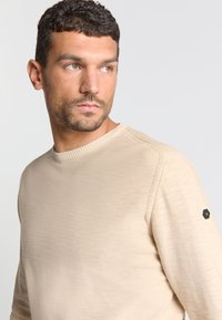 Beige ronde hals trui gemaakt van zachte stof met een ribgebreide halslijn en een hexagonaal zwart logopatch op de linkermouw.