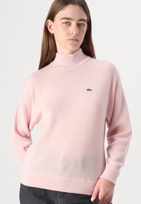 Ljusrosa polotröja i mjukt, texturerat stickat material. Har en liten grön logotyp på bröstet. Ribbad mudd och nederkant ger struktur.