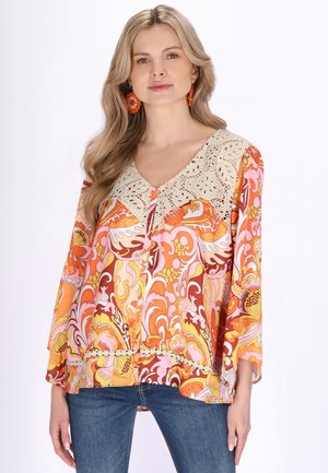 Młoda kobieta z długimi blond włosami ubrana w jasnopomarańczową bluzkę z wzorem, z koronkowym karczkiem, oraz niebieskie dżinsy, stoi na białym tle.