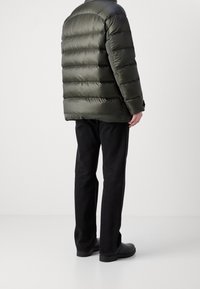 Chaqueta acolchada de color verde oliva con un diseño acolchado, combinada con pantalones negros, costuras visibles y zapatos negros, de pie sobre un fondo neutro.