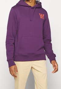 Fialová mikina s kapucí vyrobená z bavlněné směsi, s přední kapsou, nastavovací kapucí na šňůrku a oranžovým logem na hrudi.