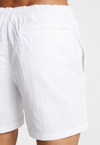 Fehér shorts rugalmas derékkal, hátul cipzáras zsebbel és oldalzsebekkel. Az anyag könnyűnek és texturáltnak tűnik.