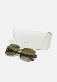 Ochelari de soare supradimensionați verzi cu lentile maronii degrade, având o ramă robustă și un toc alb gravat cu „MICHAEL KORS.”
